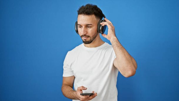 Jonge Arabier man luisteren naar muziek over geïsoleerde blauwe achtergrond - Foto, afbeelding
