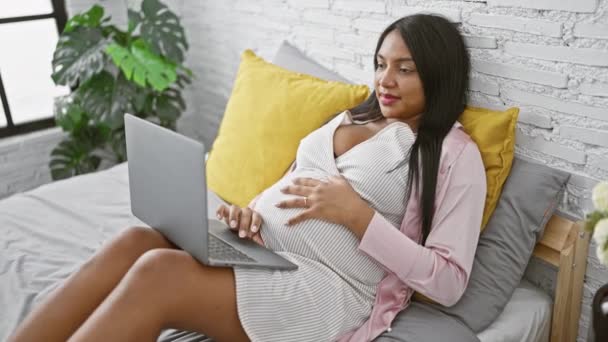 Sebevědomá mladá těhotná žena, šťastně se povalující ve své útulné ložnici, odpočívající na posteli s laptopem, dotýkající se svého břicha v klidu slunečného rána. - Záběry, video