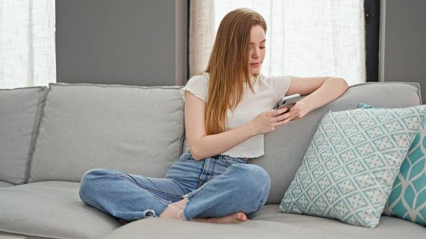 Young blonde woman using smartphone sitting on sofa at home - Photo, Image