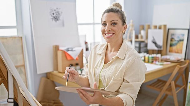 Young blonde woman artist smiling confident holding paintbrush and palette at art studio - Photo, Image