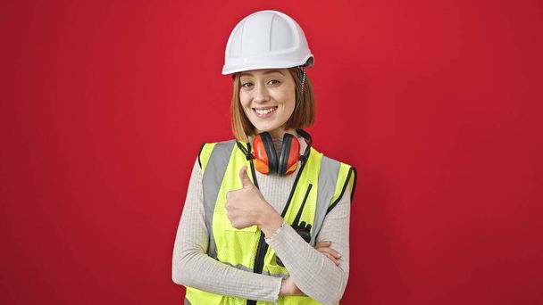 Jeune femme blonde architecte debout avec les bras croisés geste faire pouce vers le haut geste sur fond rouge isolé - Photo, image