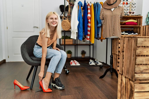 Jovem loira cliente vestindo sapatos de salto alto sentado na cadeira na loja de roupas - Foto, Imagem
