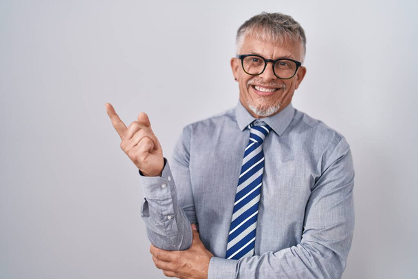 Latinalaisamerikkalainen liikemies harmaat hiukset yllään lasit iso hymy kasvoillaan, osoittaen sormella sivuun katsoen kameraa.  - Valokuva, kuva