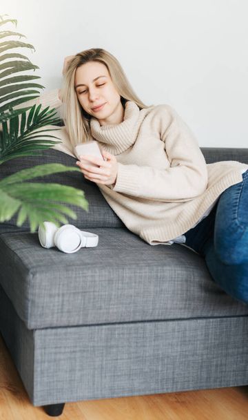 Glückliche junge Frau lächelt und benutzt Handy, während sie auf dem Sofa im Wohnzimmer sitzt - Foto, Bild