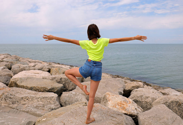 юная стройная девушка занимается гимнастическими упражнениями на скалах у моря в джинсовых шортах и футболке с коротким рукавом летом - Фото, изображение