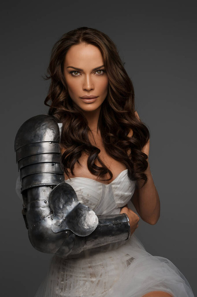 Elegant middle-aged brunette in a white gown and partial medieval armor poses on a gray backdrop - Photo, Image