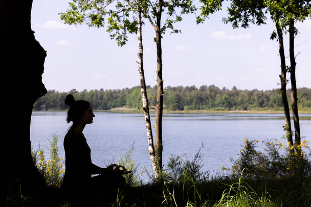 Een sereen silhouet van een jonge duizendjarige vrouw die yoga beoefent en mediteert op weelderig groen gras naast een rustig bosmeer, dat de sereniteit van de natuur omarmt. Yoga Meditatie bij Forest Lake. Hoog - Foto, afbeelding