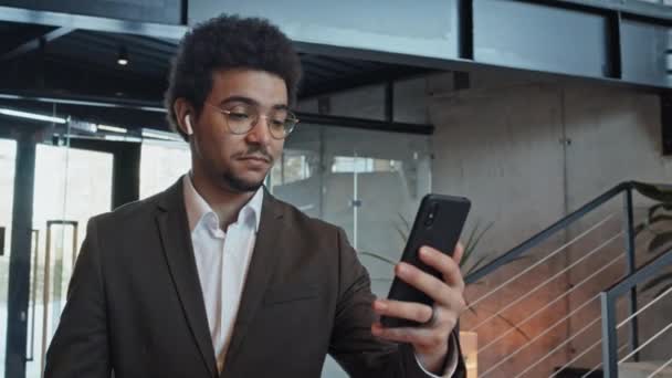 Plan rapproché moyen du jeune homme du Moyen-Orient en lunettes et costume décontracté, portant des écouteurs sans fil tenant le smartphone et parlant sur appel vidéo sur le chemin du bureau - Séquence, vidéo
