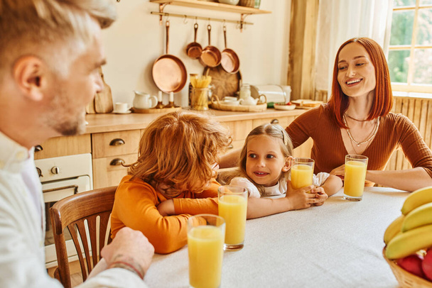 キッチンでの朝食中にオレンジジュースや果物の近くに娘と息子と陽気な両親 - 写真・画像