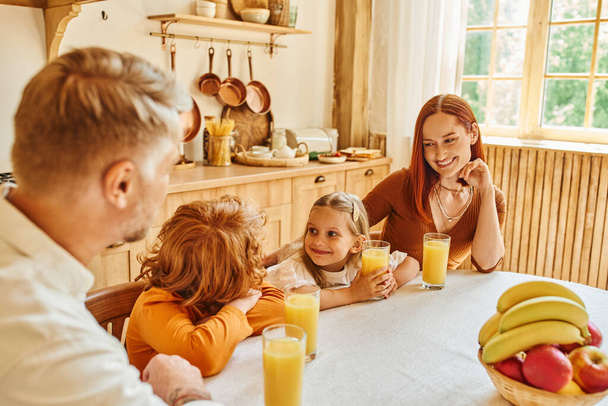 радісний брат і сестра посміхаються один одному біля свіжого апельсинового соку і батьків на кухні вдома - Фото, зображення
