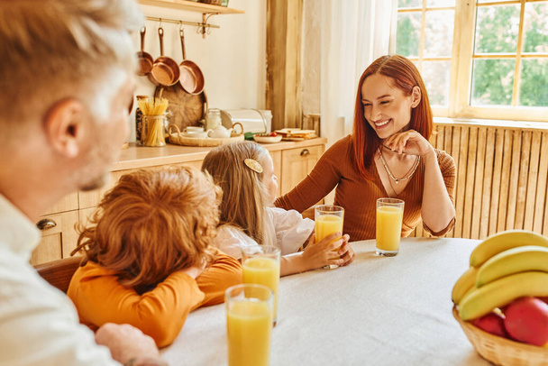 Gülümseyen bir kadın taze portakal suyunun yanındaki çocuklara bakıyor ve kahvaltıda da evde. - Fotoğraf, Görsel