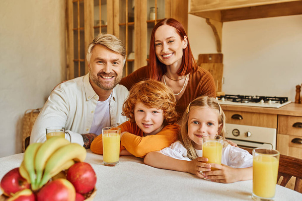 キッチンで新鮮な果物やオレンジジュースの近くでカメラを見ている愛らしい子供たちと楽しいカップル - 写真・画像