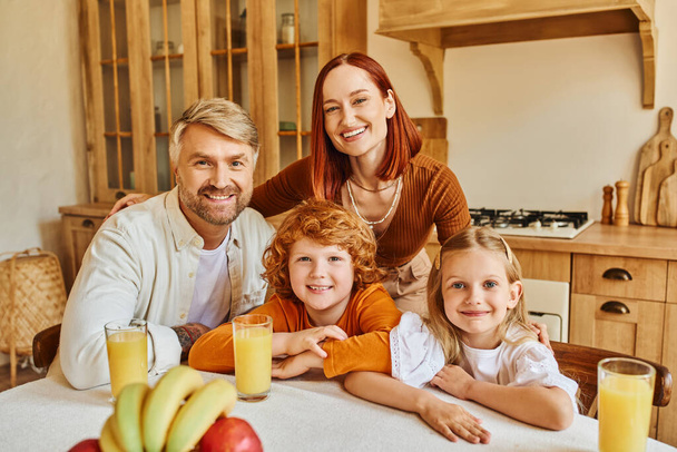 радісні батьки з чарівними дітьми дивляться на камеру біля свіжих фруктів і апельсиновий сік на кухні - Фото, зображення