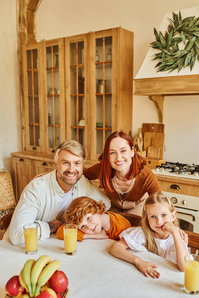 parents with cute children looking at camera near during breakfast in cozy kitchen, smiling faces - Photo, Image