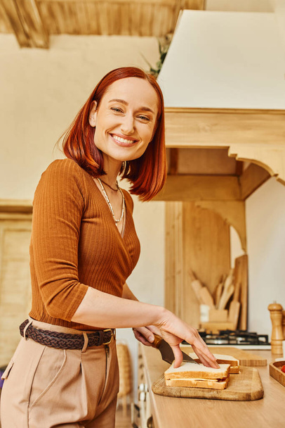 vrolijke roodharige vrouw op zoek naar camera en het bereiden van heerlijke broodjes voor het ontbijt in de keuken - Foto, afbeelding