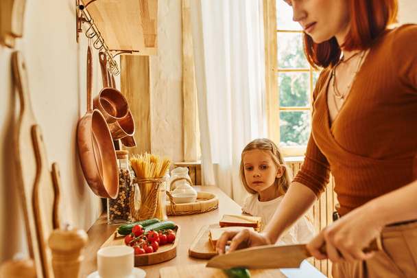 sorridente madre tagliare le verdure preparare la colazione vicino figlia con panino sul bancone della cucina - Foto, immagini