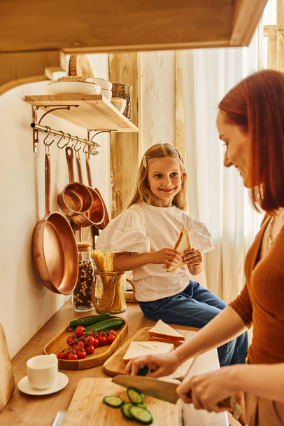 menina feliz sentada com sanduíche no balcão da cozinha perto de mãe sorridente preparando café da manhã - Foto, Imagem