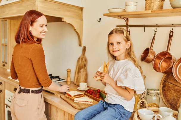 усміхнена дівчина сидить зі сніданком на кухонній стійці, а щаслива мати готує сніданок - Фото, зображення