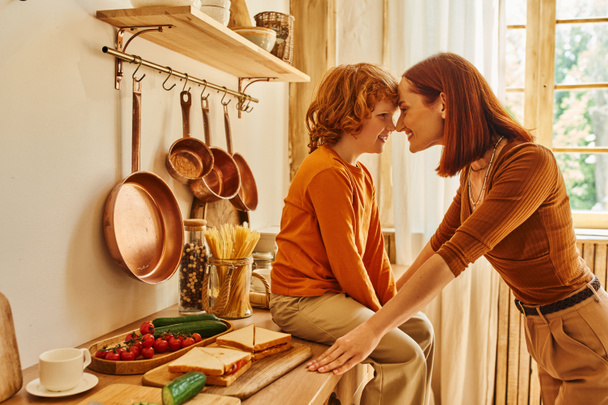 radosny dzieciak na blacie kuchennym obok kanapek i świeżych warzyw twarzą w twarz z uśmiechniętą matką - Zdjęcie, obraz