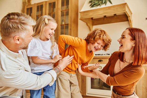 cheerful parents tickling excited kids in modern kitchen, fun and laughter in cozy home environment - Photo, Image
