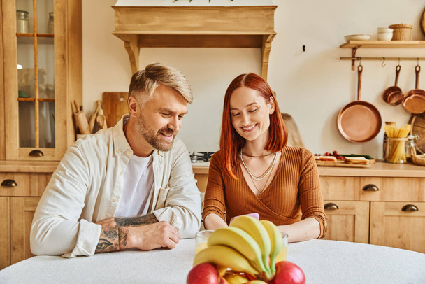 smiling woman looking at smartphone near husband and fresh fruits while sitting in modern kitchen - Photo, Image