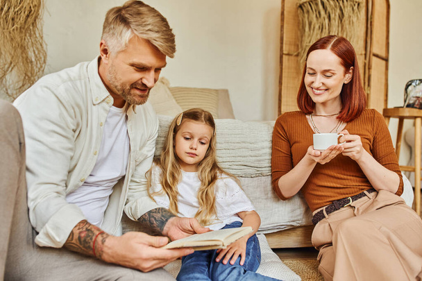 mujer sonriente con taza de café cerca de padre e hija leyendo libro en el dormitorio, aprendiendo juntos - Foto, Imagen