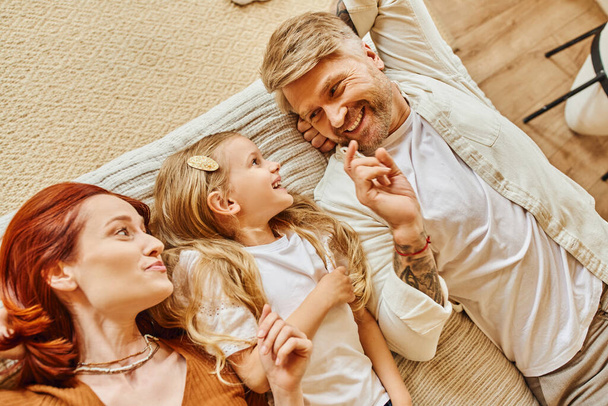 top view of cheerful parents with cute daughter lying down and talking on bed, fun and laughter - Foto, Bild