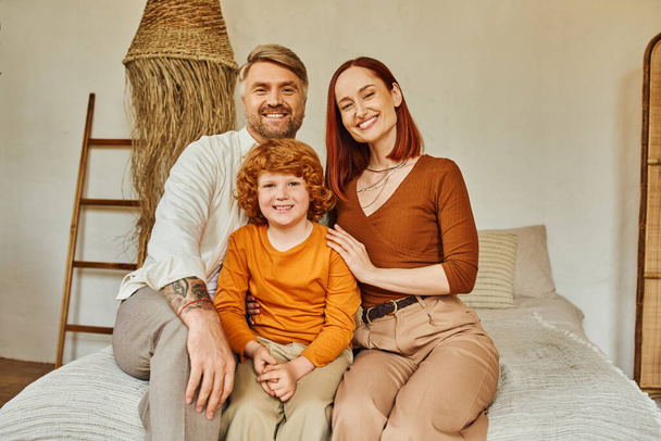 cheerful parents with redhead son sitting on bed and looking at camera in cozy home environment - Φωτογραφία, εικόνα