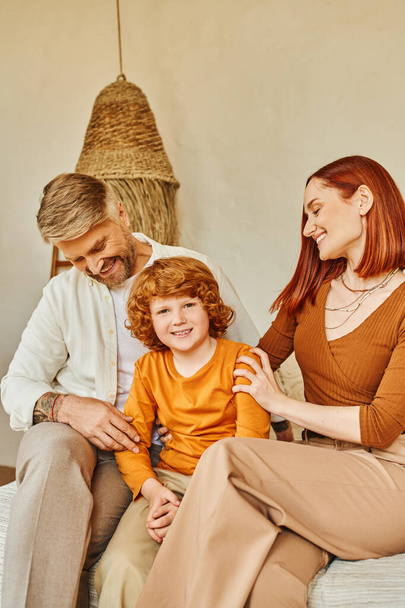 smiling husband and wife embracing redhead kid while sitting in bedroom, emotional connection - Photo, Image