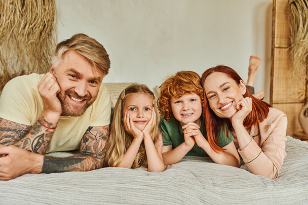 улыбающиеся родители и счастливые дети, лежащие на кровати и глядя в камеру дома, время релаксации - Фото, изображение