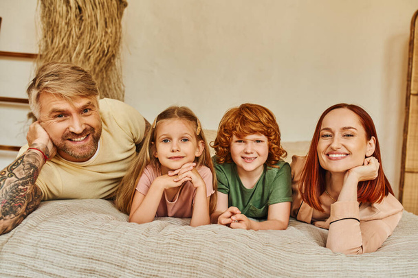 smiling parents with son and daughter lying ibn cozy bedroom and looking at camera, bonding moments - Foto, Imagem