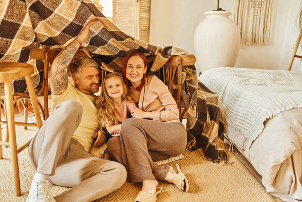 padres alegres con hija feliz escondida debajo de la cabaña manta en la sala de estar, jugando juntos - Foto, imagen