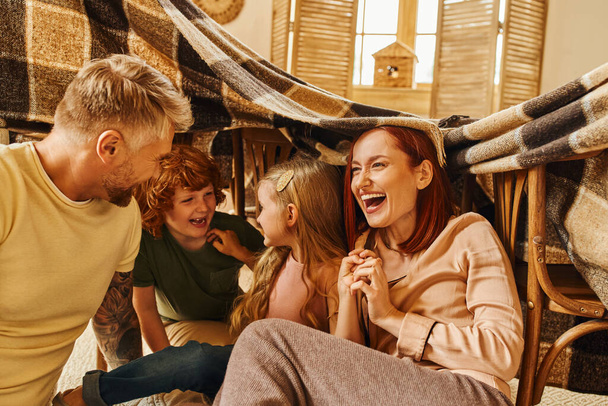 overjoyed parents laughing under blanket hut while playing with kids in living room, bonding moments - Fotó, kép