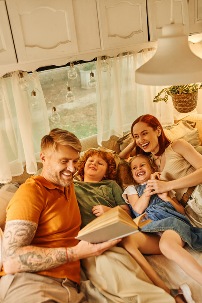 tetovált férfi olvasás könyv nevető család puha ágyon kényelmes lakókocsi otthon, tanulás együtt - Fotó, kép