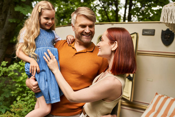 smiling man holding cute daughter near pleased wife and mobile home in trailer park, leisure - Photo, Image
