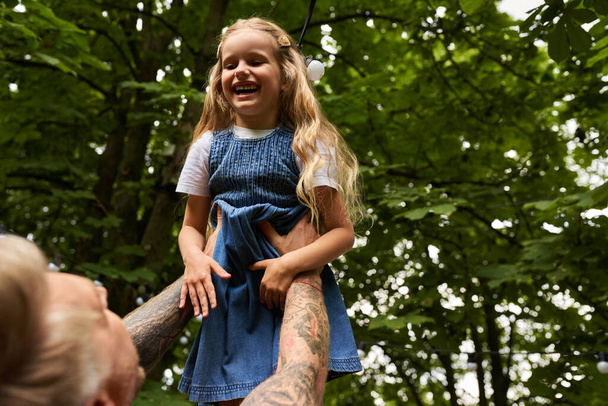 überglückliches Mädchen lacht in den Händen des tätowierten Vaters im grünen Park, Freizeit und gemeinsames Spielen - Foto, Bild