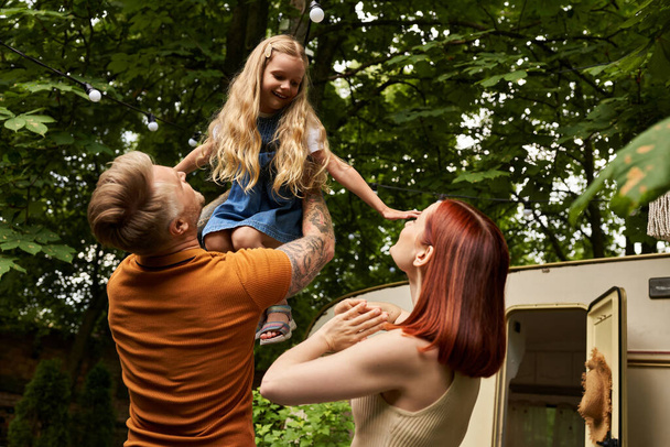tattooed man holding overjoyed daughter in hands near wife and modern trailer home, family leisure - Photo, Image