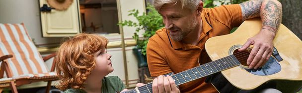 glücklicher Mann spielt Akustikgitarre vor aufmerksamem Sohn neben Wohnwagen, Spaß und Lernen, Transparent - Foto, Bild