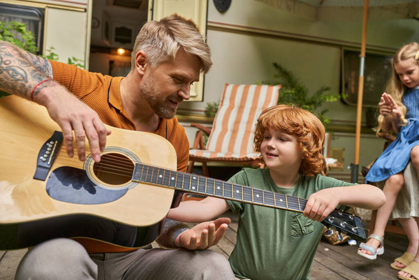 татуйований чоловік навчає рудого сина грати на акустичній гітарі біля дому на колесах у парку трейлерів - Фото, зображення