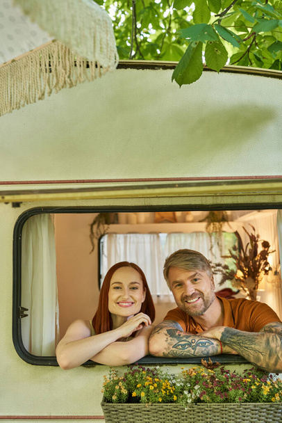 happy tattooed man and redhead woman looking out window of modern trailer home, leisure and fun - Photo, Image