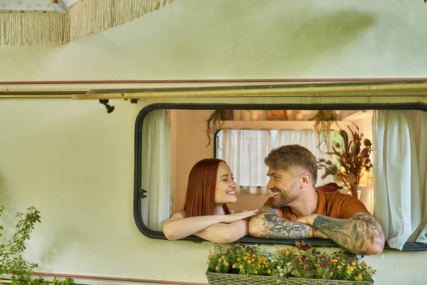 feliz tatuado homem e ruiva mulher olhando uns para os outros na janela do moderno reboque casa - Foto, Imagem