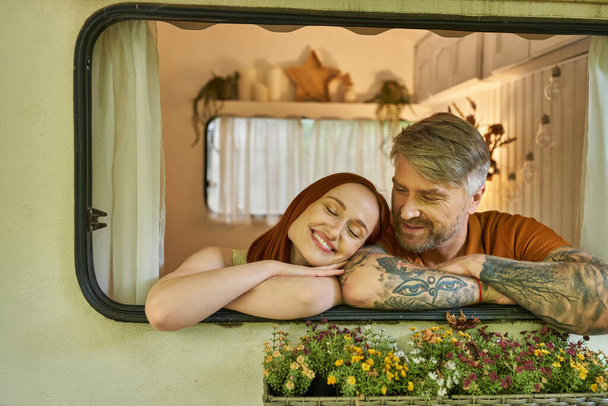 heureux rousse femme souriant avec les yeux fermés près de tatoué homme dans la fenêtre de remorque moderne maison - Photo, image
