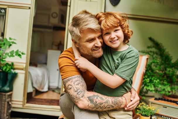 boldog tetovált férfi átöleli imádnivaló vörös hajú fia közelében modern lakókocsi otthon, kötődés pillanatok - Fotó, kép