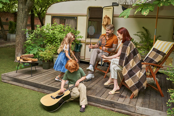 boy learning to play guitar near sister and parents with badminton rockets next to trailer home - Photo, Image