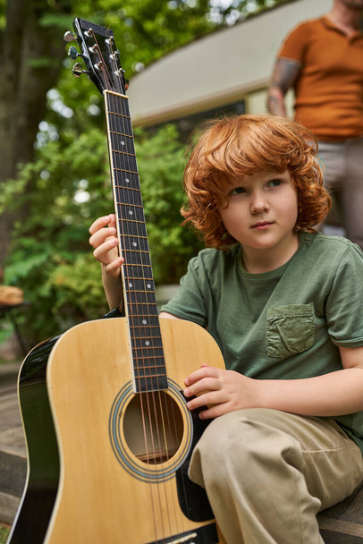 мечтательный рыжий мальчик с акустической гитарой смотрит в сторону, сидя рядом с современным трейлером - Фото, изображение