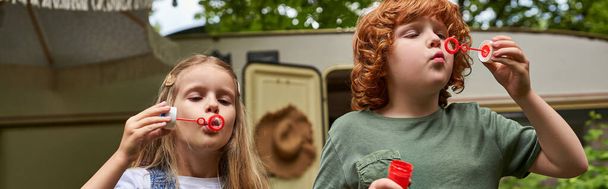 adorable children blowing soap bubbles near trailer home outdoors, siblings playing together, banner - Photo, Image