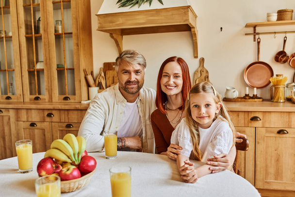 boldog szülők aranyos lánya néz kamera közelében friss gyümölcsök és narancslé a konyhában - Fotó, kép