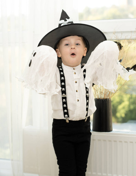 Halloween concept. Een kleine knappe jongen met een tovenaarshoed met witte geesten speelt vrolijk en emotioneel in een interieur tegen de achtergrond van een raam. Close-up. - Foto, afbeelding