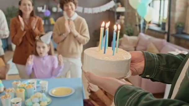 Primer plano seguimiento disparo de las manos de un hombre irreconocible llevando pastel de cumpleaños con velas encendidas y ponerlo en la mesa delante de los niños pequeños y emocionados, y los padres aplaudiendo y aplaudiendo - Imágenes, Vídeo