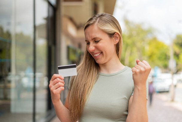 Mujer rubia joven sosteniendo una tarjeta de crédito al aire libre celebrando una victoria - Foto, Imagen